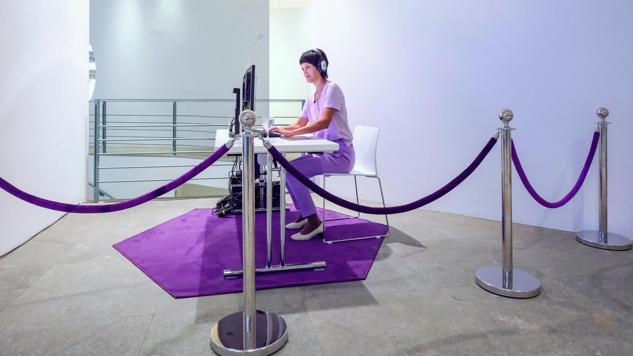 En person sitter vid ett skrivbord med hörlurar framför datorskärmar. I förgrunden syns hur rummet är avgränsas med stolpar och rep.