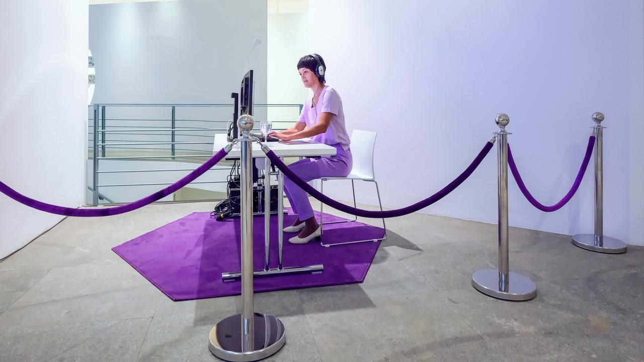 En person sitter vid ett skrivbord med hörlurar framför datorskärmar. I förgrunden syns hur rummet är avgränsas med stolpar och rep.